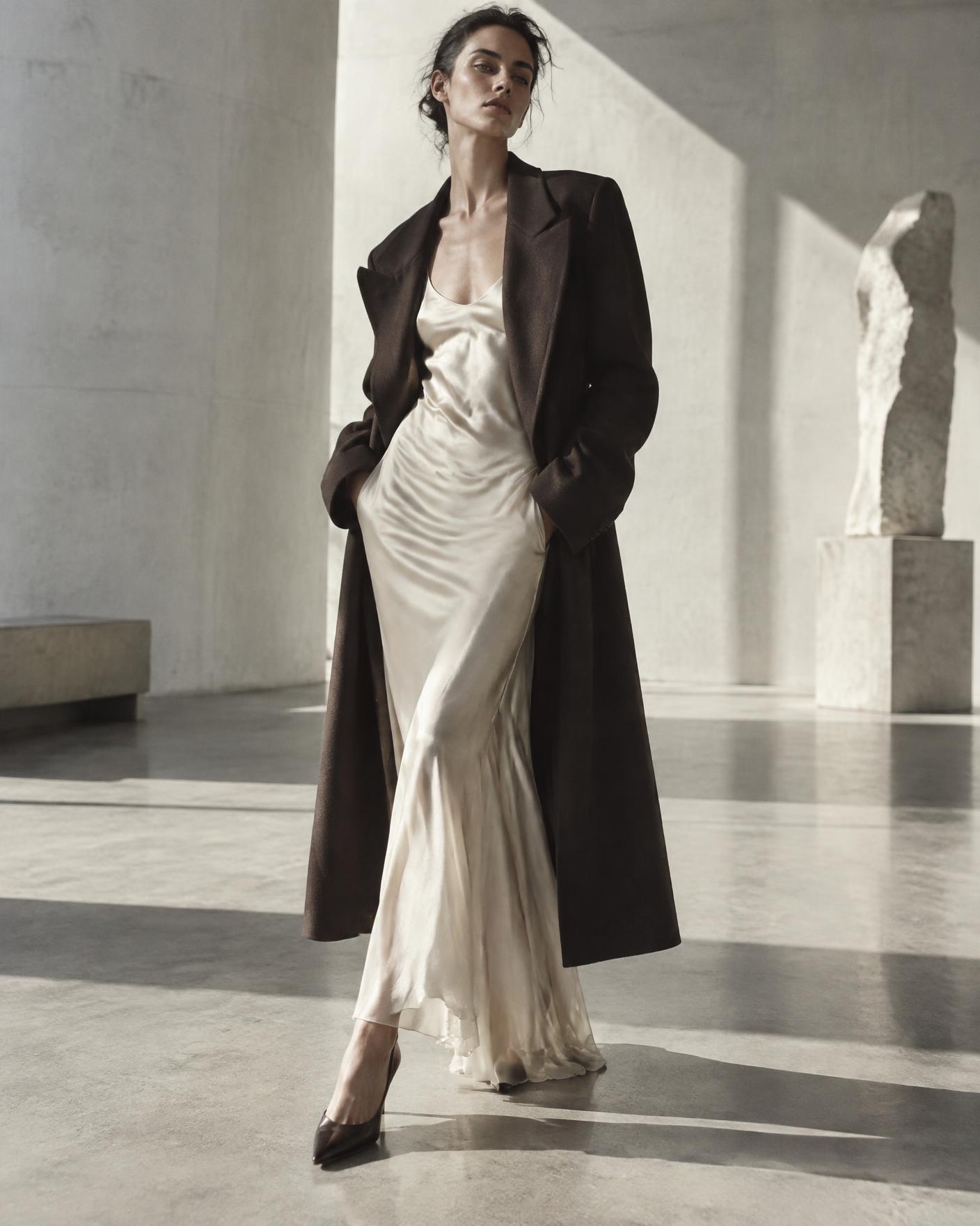 Model in an ivory silk slip dress and oversized espresso tailored coat in a bright concrete gallery